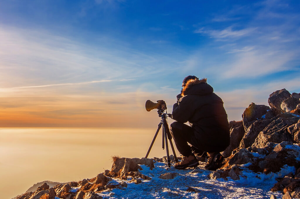 Traveller takes photos with camera on tripod on rocky peak at sunset.